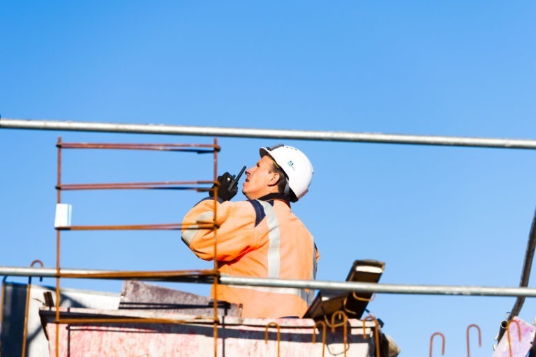 Un ouvrier du bâtiment portant un gilet orange haute visibilité et un casque de chantier blanc utilise un talkie-walkie. L'ouvrier est entouré d'échafaudages sur fond de ciel bleu clair, avec des barres métalliques et des matériaux de construction autour d'eux, représentant Delta Construction à Bordeaux sur son site actif.