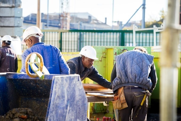 Des ouvriers du bâtiment portant des casques blancs et des gilets de protection travaillent sur un chantier de Delta Construction à Bordeaux. Un ouvrier est penché sur un engin, tandis qu'un autre manipule des matériaux sur une table. Un conteneur bleu et divers outils sont visibles. L'arrière-plan montre une zone clôturée avec des structures industrielles.