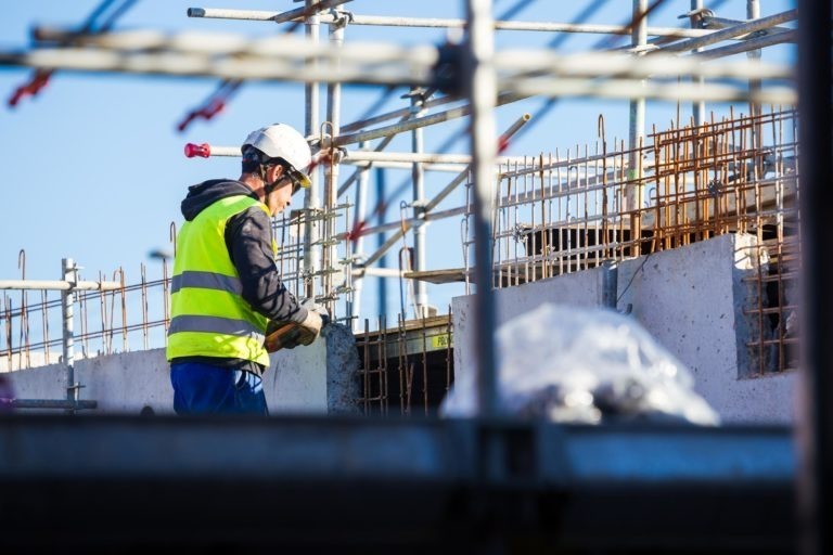 Un ouvrier du bâtiment portant un casque de sécurité blanc, un gilet de sécurité jaune et des gants travaille sur un chantier de Delta Construction à Bordeaux entouré d'échafaudages et de barres d'armature métalliques. L'ouvrier semble manipuler un équipement ou un matériau à deux mains. Le ciel est clair et bleu en arrière-plan.