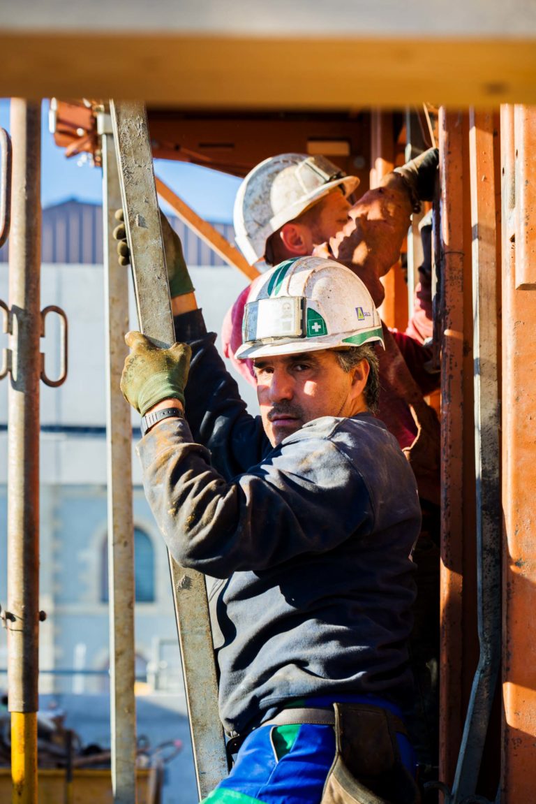 Deux ouvriers du bâtiment de Delta Construction à Bordeaux portent un casque et des gants de sécurité alors qu'ils travaillent sur un chantier industriel. L'ouvrier au premier plan tient une poutre métallique et regarde la caméra, tandis que l'ouvrier à l'arrière-plan ajuste un équipement fixé à une structure.