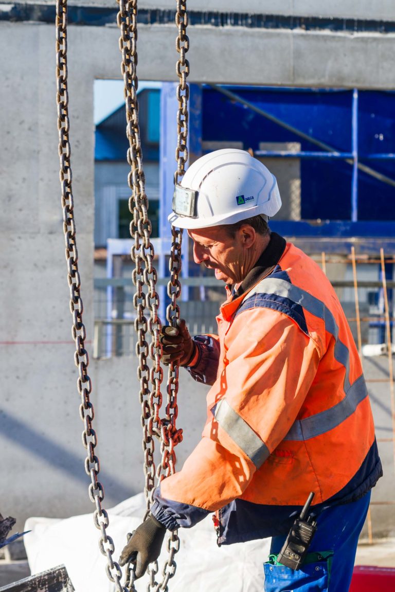 Un ouvrier du bâtiment portant un gilet orange haute visibilité et un casque de chantier blanc manœuvre de lourdes chaînes sur un chantier de Delta Construction à Bordeaux. L'ouvrier porte des gants et utilise ses deux mains pour manipuler les chaînes, avec des structures en béton et des matériaux de construction visibles en arrière-plan.