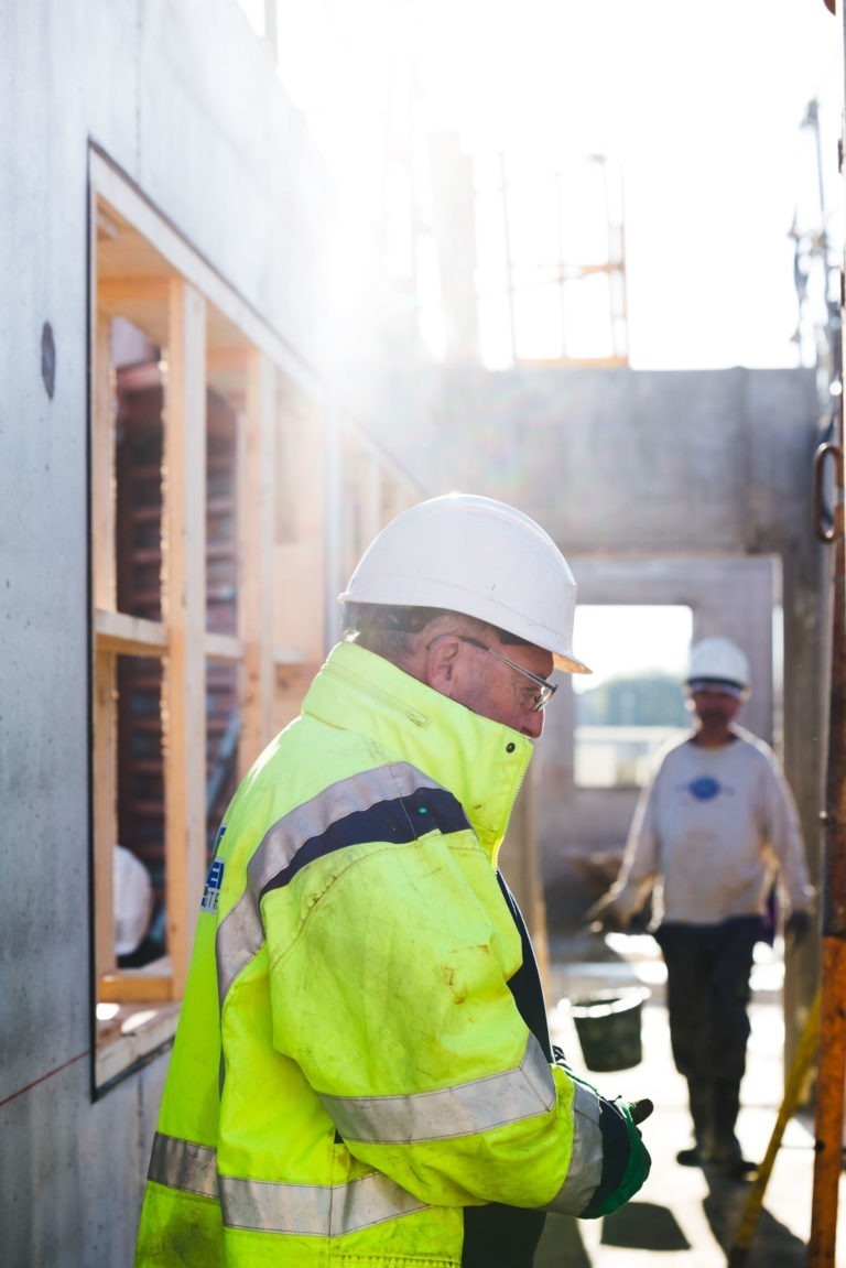 Un ouvrier du bâtiment de Delta Construction à Bordeaux, portant un casque de sécurité blanc et un gilet de haute visibilité jaune, regarde vers le bas alors qu'il se tient près d'un bâtiment en construction. Un autre ouvrier à l'arrière-plan, également portant un casque de sécurité blanc, est partiellement visible en train de marcher sur le chantier alors que la lumière du soleil brille à travers la zone.