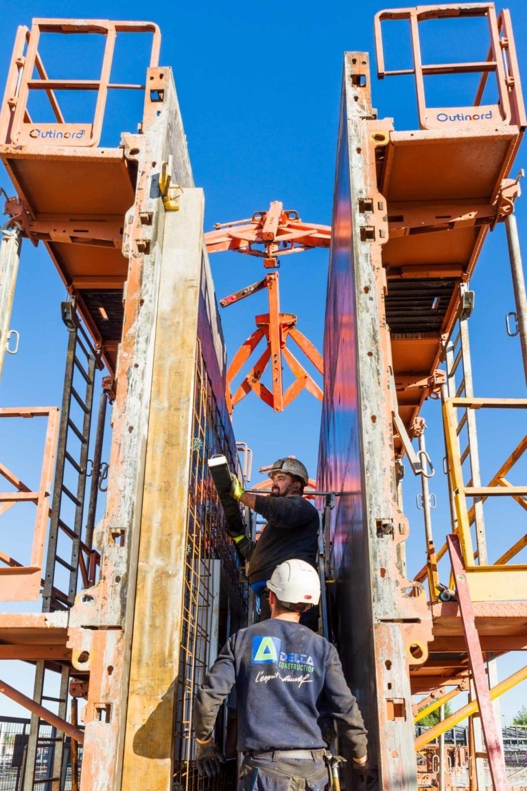 Deux ouvriers de Delta Construction à Bordeaux portent des casques de sécurité et des gilets de haute visibilité. Ils travaillent avec de grands moules en béton verticaux, orange et gris. L'un des ouvriers se tient debout sur un échafaudage et fixe le moule avec un outil, tandis que l'autre observe. Le ciel est clair et bleu.