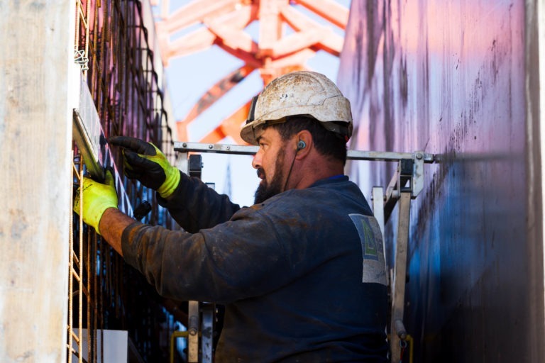 Un ouvrier du bâtiment portant un casque de sécurité et des protections auditives installe des barres d'armature en acier dans une étroite structure en béton chez Delta Construction à Bordeaux. Il porte un sweat-shirt sombre et des gants jaune vif, travaillant avec une expression concentrée. Une structure est visible en arrière-plan sous un ciel lumineux.