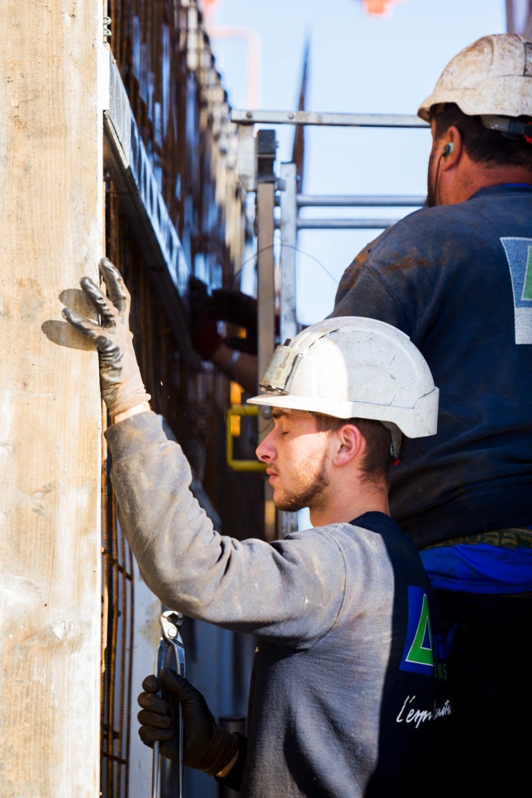 Deux ouvriers du bâtiment portant des casques de sécurité blancs et des équipements de sécurité bleus travaillent avec diligence sur un chantier de Delta Construction à Bordeaux. L'un tient une clé à molette et positionne un cadre métallique, tandis que l'autre se tient derrière et observe. Tous deux portent des gants noirs et restent concentrés sous la lumière vive du soleil.