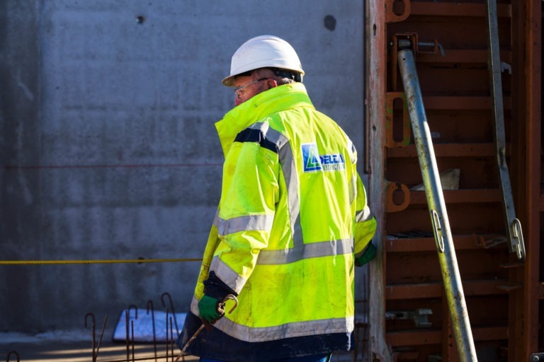 Un ouvrier du bâtiment portant un gilet de haute visibilité jaune vif et un casque blanc se tient près d'un échafaudage métallique sur le chantier de Delta Construction à Bordeaux. L'ouvrier regarde dans le vide, concentré sur le chantier. L'arrière-plan montre un mur en béton et divers matériaux de construction, avec des barres d'armature visibles au premier plan.