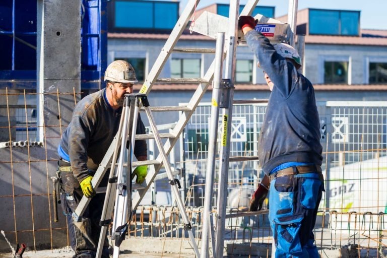 Deux ouvriers du bâtiment en tenue de sécurité de Delta Construction à Bordeaux collaborent pour positionner une charpente métallique sur un chantier. L'un soutient un escabeau tandis que l'autre ajuste la structure. Ils sont entourés de clôtures et de murs en béton partiellement construits, avec des bâtiments visibles en arrière-plan.