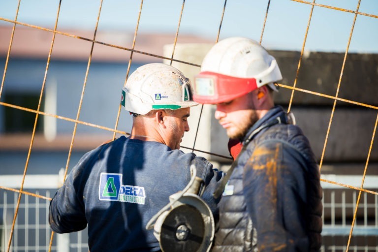 Deux ouvriers du bâtiment de Delta Construction à Bordeaux, coiffés de casques et d'équipements de protection, travaillent sur un chantier. L'un, vêtu d'une chemise bleue avec logo et d'un casque blanc, fixe une grille métallique. L'autre, vêtu d'une veste et d'un casque rouge, regarde vers le bas en tenant un outil. Tous deux semblent concentrés au milieu des matériaux de construction.