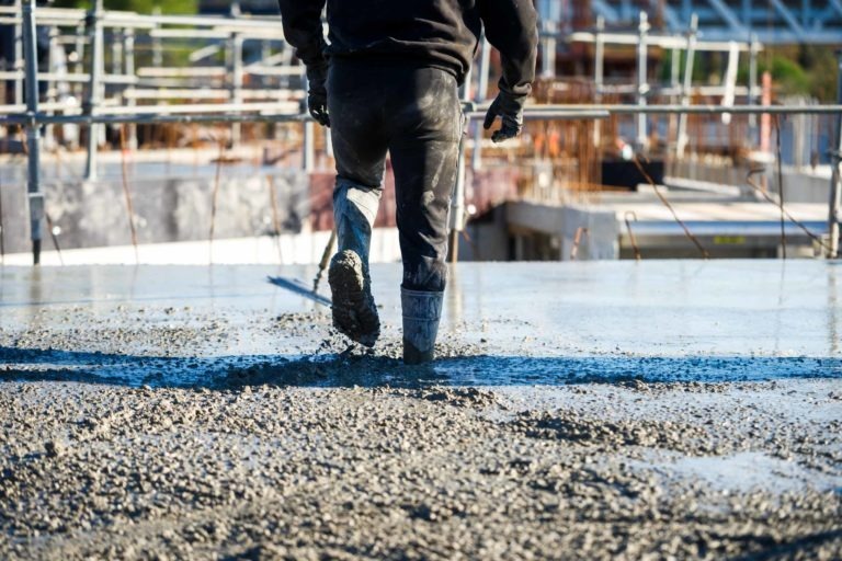 Un ouvrier de Delta Construction à Bordeaux marche sur le béton humide d'un chantier de construction. L'ouvrier porte des vêtements noirs, de hautes bottes en caoutchouc et des gants. L'arrière-plan présente des échafaudages, des barres d'acier et d'autres éléments de construction sous un ciel dégagé. L'identité de l'ouvrier n'est pas visible.