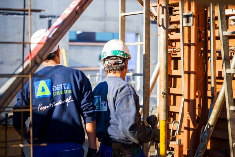 Deux ouvriers du bâtiment en uniforme bleu et casque blanc se tiennent devant un échafaudage. L'un d'eux tient un outil jaune. Leurs uniformes arborent le logo de Delta Construction à Bordeaux et le texte « lambdaTeam plus ». L'arrière-plan représente un chantier de construction en extérieur avec des structures en béton.