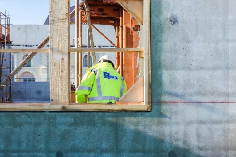 Un ouvrier du bâtiment portant un gilet de sécurité et un casque de chantier, arborant le logo « Delta Construction à Bordeaux », est aperçu à travers le cadre d'une fenêtre d'un bâtiment en béton inachevé. La charpente et l'échafaudage du bâtiment sont visibles, suggérant que des travaux de construction sont en cours.