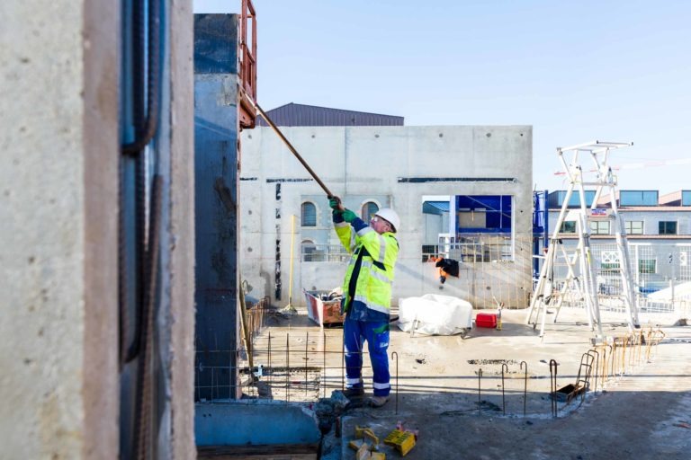 Un ouvrier du bâtiment portant une veste jaune haute visibilité et un casque de chantier, représentant Delta Construction à Bordeaux, utilise un long outil, peut-être un rouleau à peinture, sur un haut mur de béton. Autour du travailleur se trouvent divers matériaux de construction, des barres d'armature et des équipements, avec des structures en béton non finies et des échafaudages en arrière-plan.