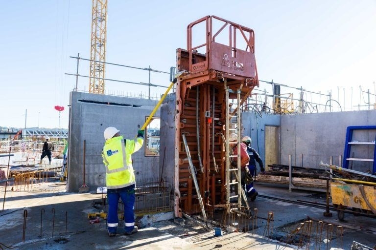 Des ouvriers du bâtiment de Delta Construction à Bordeaux sont sur un chantier. Un ouvrier portant un gilet jaune haute visibilité et un casque blanc utilise un outil de mesure sur un cadre métallique. Un autre ouvrier portant un gilet bleu et un casque blanc se trouve à proximité. La zone est entourée de murs en béton, de barres d'armature et d'engins de chantier.