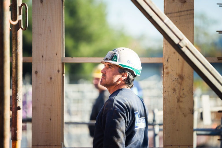 Un ouvrier du bâtiment portant un casque de chantier Delta Construction à Bordeaux et un sweat-shirt foncé regarde vers le haut alors qu'il se tient sur un chantier. Une charpente en bois et des éléments métalliques l'entourent, avec de la verdure floue et d'autres ouvriers visibles en arrière-plan, indiquant un travail actif en cours.