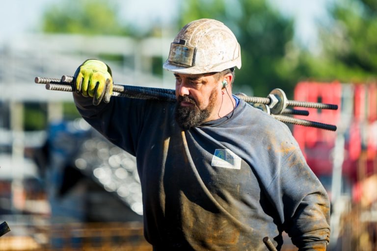 Un ouvrier du bâtiment avec une barbe et un casque sale, représentant Delta Construction à Bordeaux, porte plusieurs tiges de fer sur son épaule. Il porte une chemise à manches longues sale et des gants verts. L'arrière-plan montre des matériaux et des équipements de construction flous, avec des arbres et un ciel bleu visibles au loin.