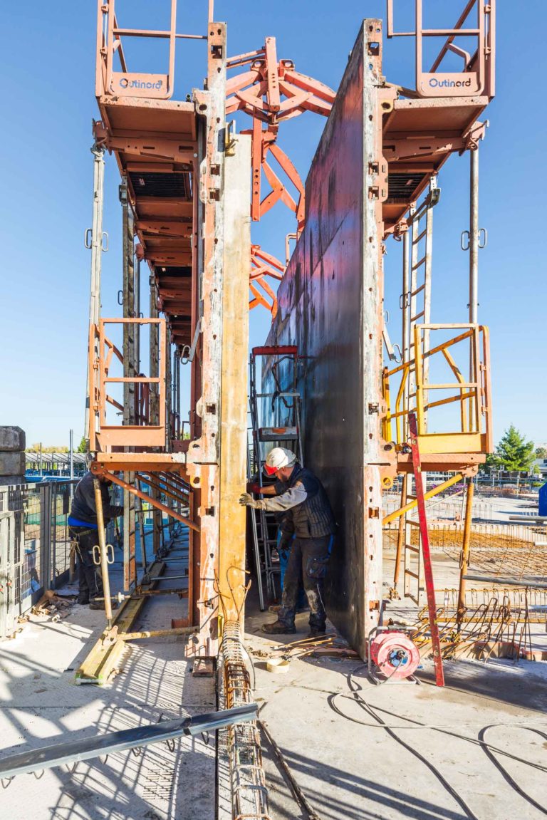 Un ouvrier de Delta Construction à Bordeaux portant un casque et un gilet de sécurité travaille entre de hautes structures de coffrage en béton verticales, entouré d'échafaudages et d'échelles. Le chantier est sous la lumière du jour, avec divers équipements de construction et des barres d'armature visibles sur le sol autour d'eux.