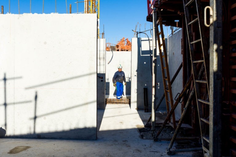 Un ouvrier du bâtiment portant un casque de sécurité, une chemise bleue et un pantalon bleu marche dans une structure en béton partiellement construite avec des barres d'armature et des échafaudages exposés sous un ciel bleu clair. Les ombres de la structure et de l'équipement sont projetées sur le sol, indiquant la lumière du soleil, sur le chantier de Delta Construction à Bordeaux.