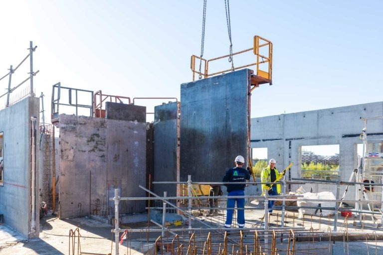 Sur un chantier, deux ouvriers en tenue de protection et casque, représentant Delta Construction à Bordeaux, guident un grand panneau de mur en béton soulevé par une grue. Des tiges d'acier et des échafaudages sont visibles, avec des structures partiellement construites en arrière-plan sous un ciel dégagé.