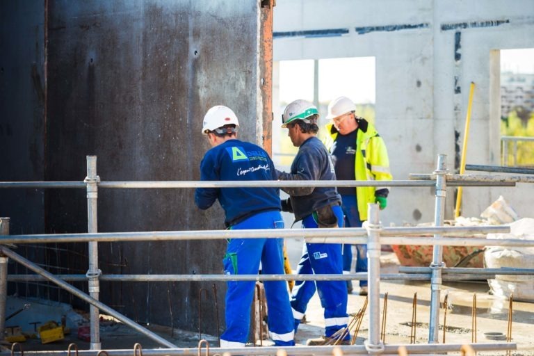 Trois ouvriers du bâtiment en uniforme bleu et casque blanc travaillent sur la structure d'un bâtiment. Deux se concentrent sur un panneau métallique, tandis qu'un autre, vêtu d'une veste jaune, les supervise. Le chantier de Delta Construction à Bordeaux comporte des murs en béton partiellement construits, des garde-corps métalliques et des matériaux éparpillés un peu partout.