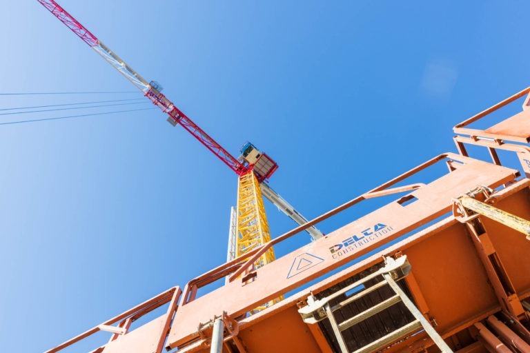 Une grue de chantier se dresse sur un ciel bleu clair. La grue, dotée d'une structure jaune et rouge, s'élève du sol à Bordeaux. Au premier plan, on peut voir une partie d'une plate-forme de chantier orange sur laquelle est écrit "Delta Construction". La photo capture une perspective ascendante.