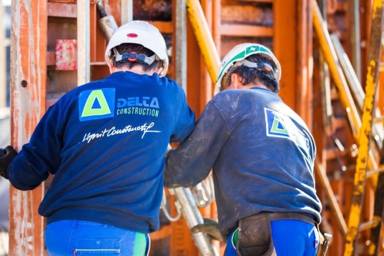 Deux ouvriers du bâtiment portant des casques et des sweat-shirts bleus avec le logo « Delta Construction à Bordeaux » travaillent en étroite collaboration sur une structure d'échafaudage orange. L'un des ouvriers tient un outil, tandis que l'autre semble aider. L'arrière-plan est constitué de matériaux de construction, mettant l'accent sur un chantier en activité.