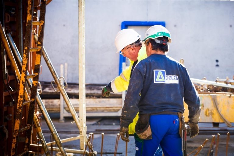 Deux ouvriers du bâtiment de Delta Construction à Bordeaux, équipés de leurs équipements de sécurité, casques et gilets de haute visibilité, collaborent sur un chantier. L'un pointe du doigt tandis que l'autre écoute. Ils sont entourés d'échafaudages et d'équipements métalliques. Le chantier a un mur en béton en arrière-plan avec une ouverture encadrée de bleu.