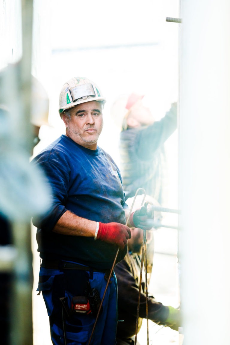 Un ouvrier du bâtiment en chemise bleue et casque de sécurité tient debout des câbles métalliques. Il porte des gants rouges et porte divers outils attachés à sa ceinture. D'autres ouvriers de Delta Construction à Bordeaux sont en arrière-plan, légèrement flous, occupés à manipuler et à installer des matériaux de construction. Ils sont entourés d'une lumière vive.