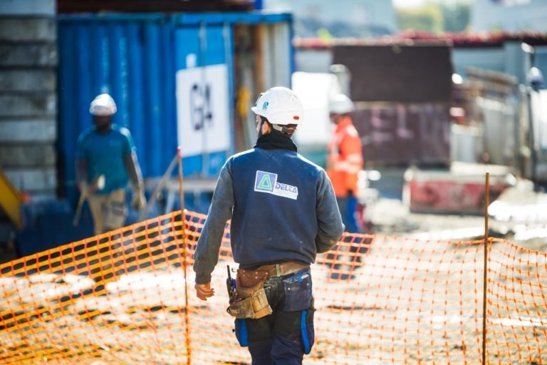 Un ouvrier du bâtiment, vêtu d'un casque de sécurité, d'un gilet de sécurité et d'une ceinture à outils, se tient debout près d'un filet de sécurité orange. Le travailleur est dos à la caméra, affichant un logo Delta Construction à Bordeaux sur sa veste. En arrière-plan, d'autres ouvriers et des matériaux de construction, dont un conteneur d'expédition bleu, sont visibles.