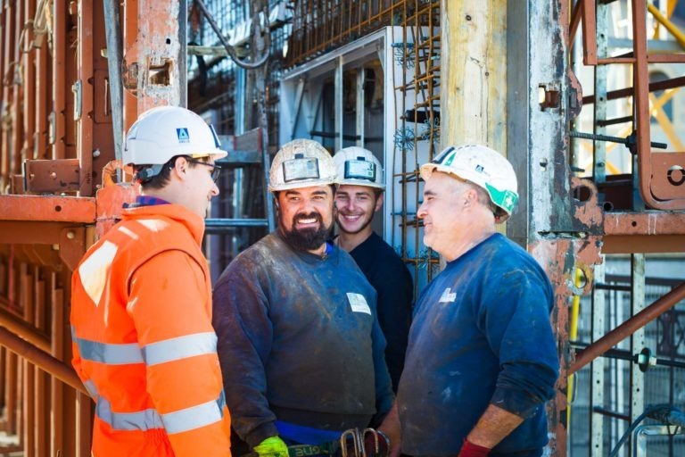 Quatre ouvriers du bâtiment de Delta Construction à Bordeaux se tiennent ensemble sur un chantier, en pleine conversation. Ils portent des casques, des équipements de sécurité et des vêtements de travail. Un ouvrier portant une veste de sécurité orange vif fait face aux trois autres, qui sourient et semblent détendus. L'arrière-plan montre des échafaudages et des équipements métalliques.