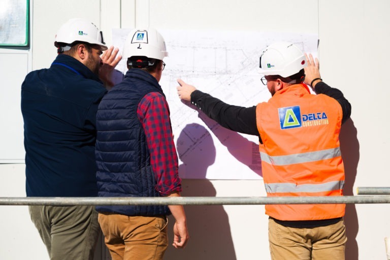 Trois ouvriers du bâtiment portant des casques de chantier examinent un plan fixé sur un mur. Un ouvrier en gilet orange pointe le plan tandis que les autres observent. Les ouvriers, qui font partie de Delta Construction à Bordeaux, sont vus de dos, tandis que la lumière du soleil projette des ombres et qu'une balustrade s'étend horizontalement au premier plan.