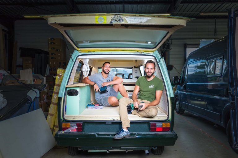 Deux hommes sont assis à l'arrière d'un camping-car ouvert dans un garage. L'un d'eux, en chemise grise, est assis sur le bord gauche, souriant, son bras reposant sur un banc en bois. L'homme de droite, en chemise verte et en tailleur, sourit tout en tenant une bouteille d'eau. Derrière eux, des cartons et un autre van, une scène authentique digne des portraits des artisans Bordeaux et Gir