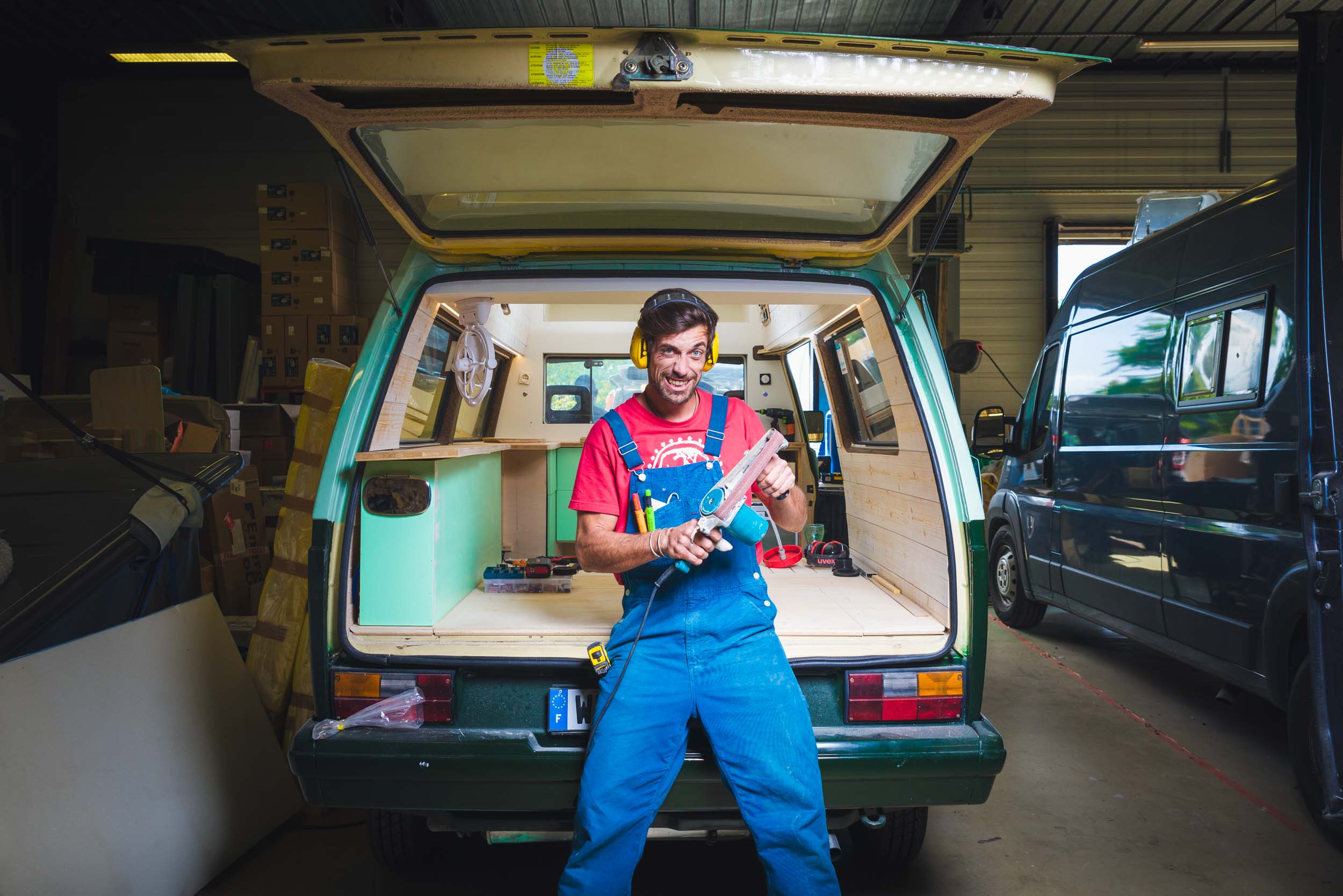 Un homme portant une salopette bleue et un t-shirt rouge se tient à l'arrière ouvert d'une camionnette verte rétro, tenant une perceuse à deux mains. L'intérieur de la camionnette est en cours de rénovation avec des panneaux en bois. L'arrière-plan présente divers outils, des boîtes et une autre camionnette à l'intérieur d'un garage à Bordeaux et Gironde. Il a une expression joyeuse.