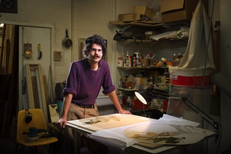 Une personne aux cheveux bouclés, à la moustache et aux lunettes se tient debout, appuyée sur une table couverte de fournitures artistiques et de portraits partiellement terminés. Ils se trouvent dans un atelier d'art rempli de divers outils, d'étagères avec des boîtes et du matériel, et d'une lampe de bureau éclairant leur espace de travail, un havre de paix pour les vrais artisans.