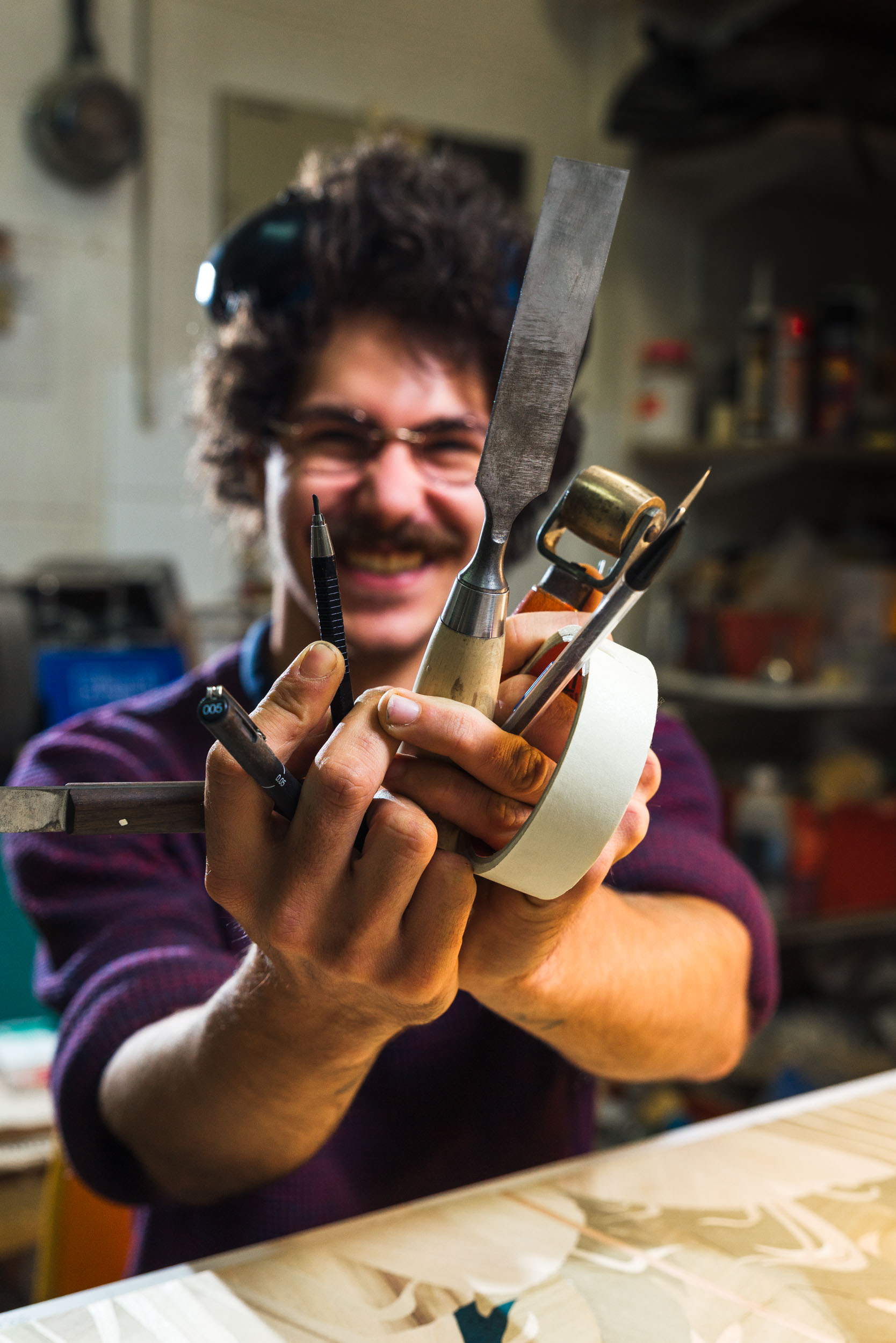 Une personne aux cheveux bouclés et aux lunettes, vêtue d'un pull sombre, sourit et tient à deux mains divers outils de travail du bois, dont des ciseaux, un petit rabot, du ruban adhésif et un crayon. Ils se trouvent dans un atelier avec des étagères et des outils en arrière-plan flou. Ce portrait capture l'essence des artisans bordelais au travail.