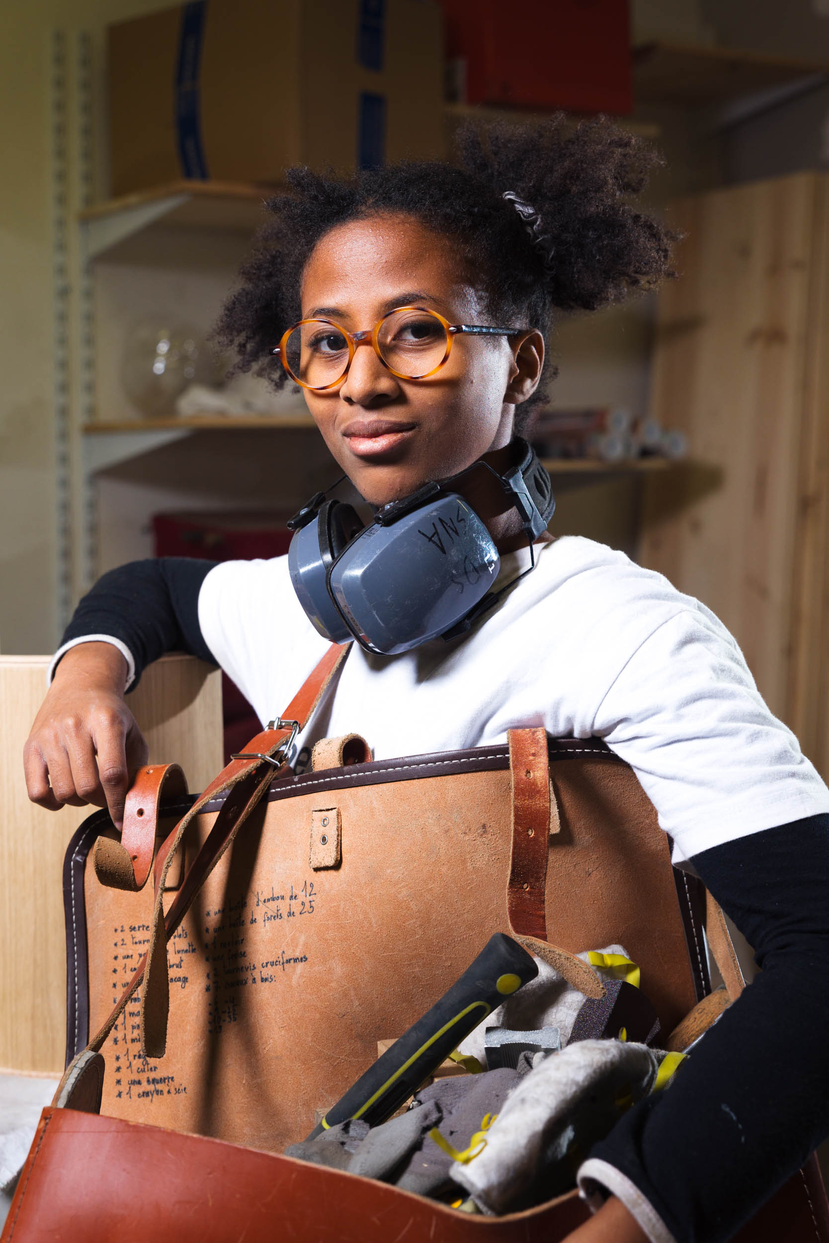 Une personne aux cheveux courts et bouclés et portant des lunettes se tient debout dans un atelier, tenant une sacoche à outils en cuir ouverte remplie de divers outils, dont des gants et des outils de coupe. Elle porte une chemise blanche avec des lunettes de sécurité autour du cou. Des étagères et des meubles en bois sont visibles en arrière-plan, capturant l'essence des artisans de Bordeaux et de Gironde.