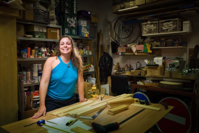 Une femme aux cheveux longs et bouclés, vêtue d'un haut bleu sans manches, sourit en s'appuyant sur un établi dans un atelier encombré. L'établi contient des planches de bois, des outils et des papiers. Les étagères à l'arrière-plan sont remplies de fournitures diverses comme des pots de peinture, des boîtes et des outils - un portrait d'artisan qui capture l'esprit de Bordeaux et de la Gironde.