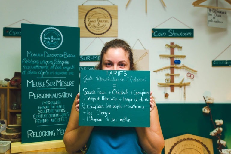 Une femme aux cheveux bruns tient un tableau noir vert sur lequel est écrit à la main le texte français « Tarifs » dans un atelier regorgeant d'objets en bois, d'étagères et de panneaux liés au mobilier et à la décoration. Cette scène captivante capture l'essence des artisans de Bordeaux et de Gironde alors qu'elle se tient au centre de la scène, cachant partiellement son visage.