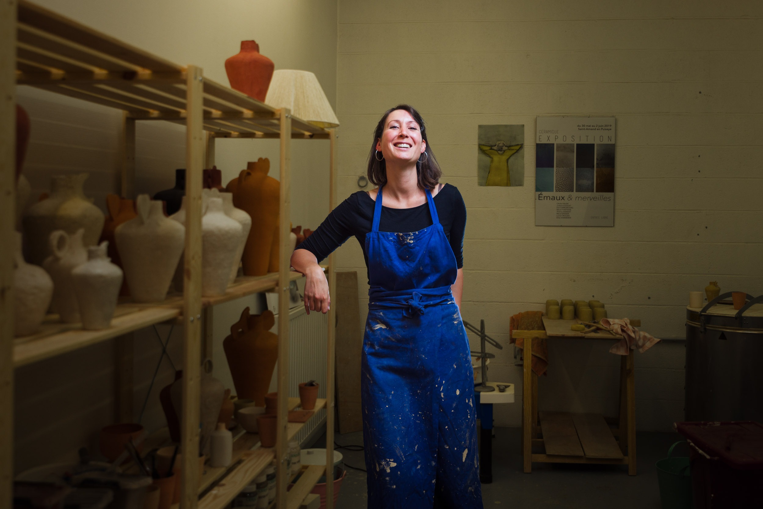 Une personne se tient debout dans un atelier de poterie, souriante et appuyée contre une étagère en bois remplie de divers vases en céramique. Elle porte un tablier bleu sur une chemise noire. La pièce, qui rappelle les ateliers d'artisans animés de Bordeaux et de Gironde, a des murs blancs avec des affiches, un four et d'autres pièces de poterie en arrière-plan. L'atmosphère est chaleureuse et créative.