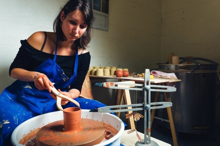 Une femme vêtue d'un haut noir et d'un tablier bleu est assise devant un tour de potier dans son atelier de Bordeaux et Gironde, façonnant l'argile avec un outil en bois. L'espace est rempli de divers outils de poterie, d'équipements et de plusieurs pièces finies sur une table. Des murs simples et de couleur claire offrent une toile de fond sereine au portrait de cette artisane.