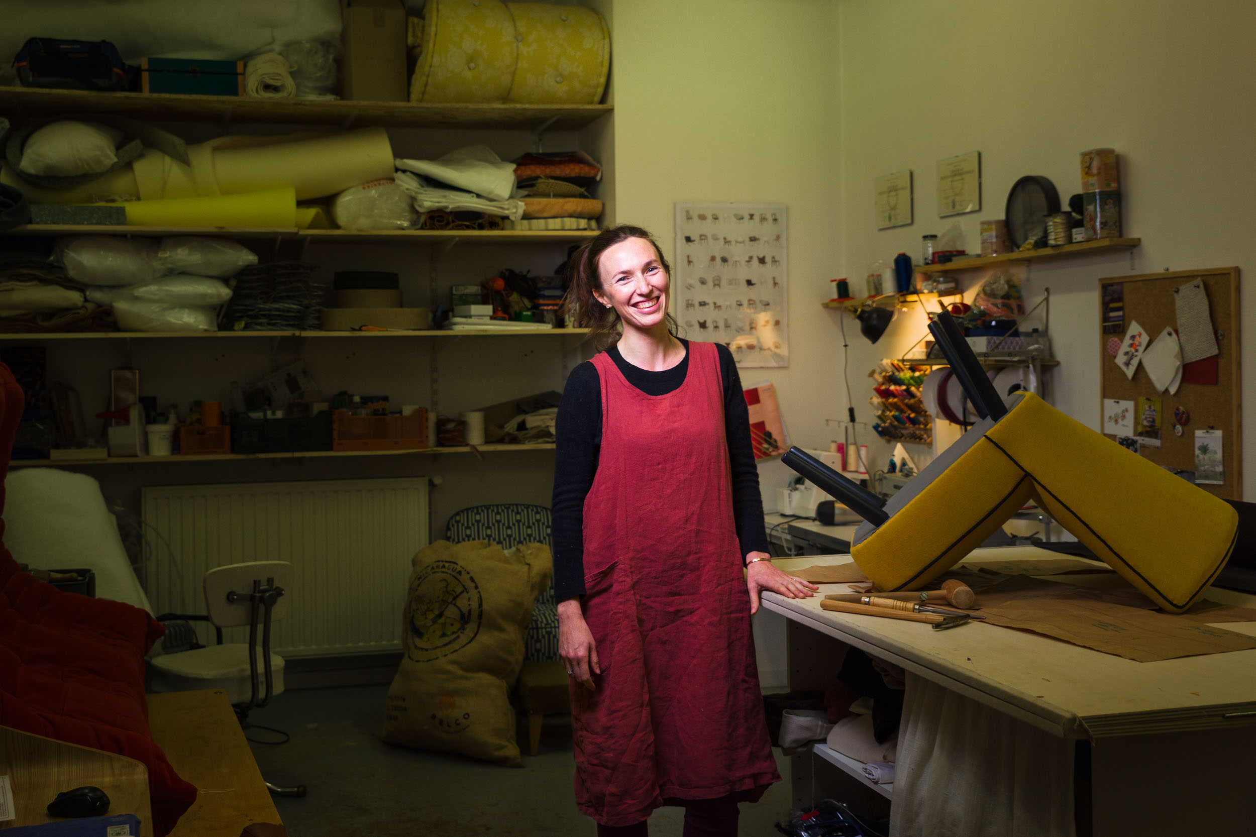 Une femme en tablier rouge se tient debout, souriante, dans un atelier rempli de divers outils et matériels de couture. Des étagères à l'arrière-plan contiennent des rouleaux de tissu et d'autres articles. Une chaise tapissée jaune est posée sur un établi au premier plan au milieu d'outils éparpillés, suggérant un projet de tapisserie réalisé par l'un des talentueux artisans portraitistes de Bordeaux et Gironde.