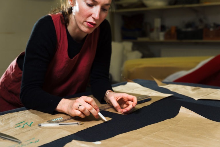 Dans un espace de travail encombré de matériaux et d'objets divers, une femme en haut noir et tablier rouge découpe avec attention un morceau de tissu à l'aide d'un petit couteau. Entourée d'outils et de patrons en papier, elle incarne les artisans concentrés souvent représentés dans les portraits de Bordeaux et de Gironde.