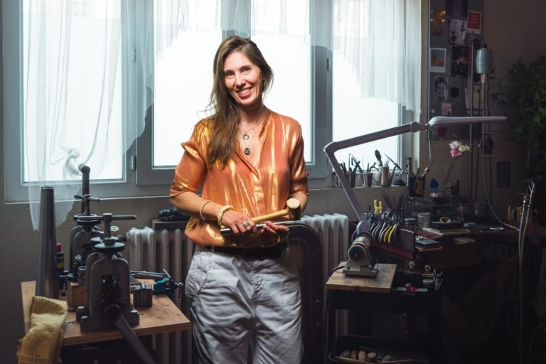 Une femme vêtue d'un chemisier doré brillant et d'un pantalon blanc se tient debout dans un atelier bien éclairé, tenant des tubes en laiton. Elle sourit et est entourée de divers outils et machines. Une grande fenêtre avec des rideaux transparents fournit de la lumière naturelle. L'espace de travail encombré respire le charme que l'on retrouve dans les portraits d'artisans de Bordeaux et de Gironde.