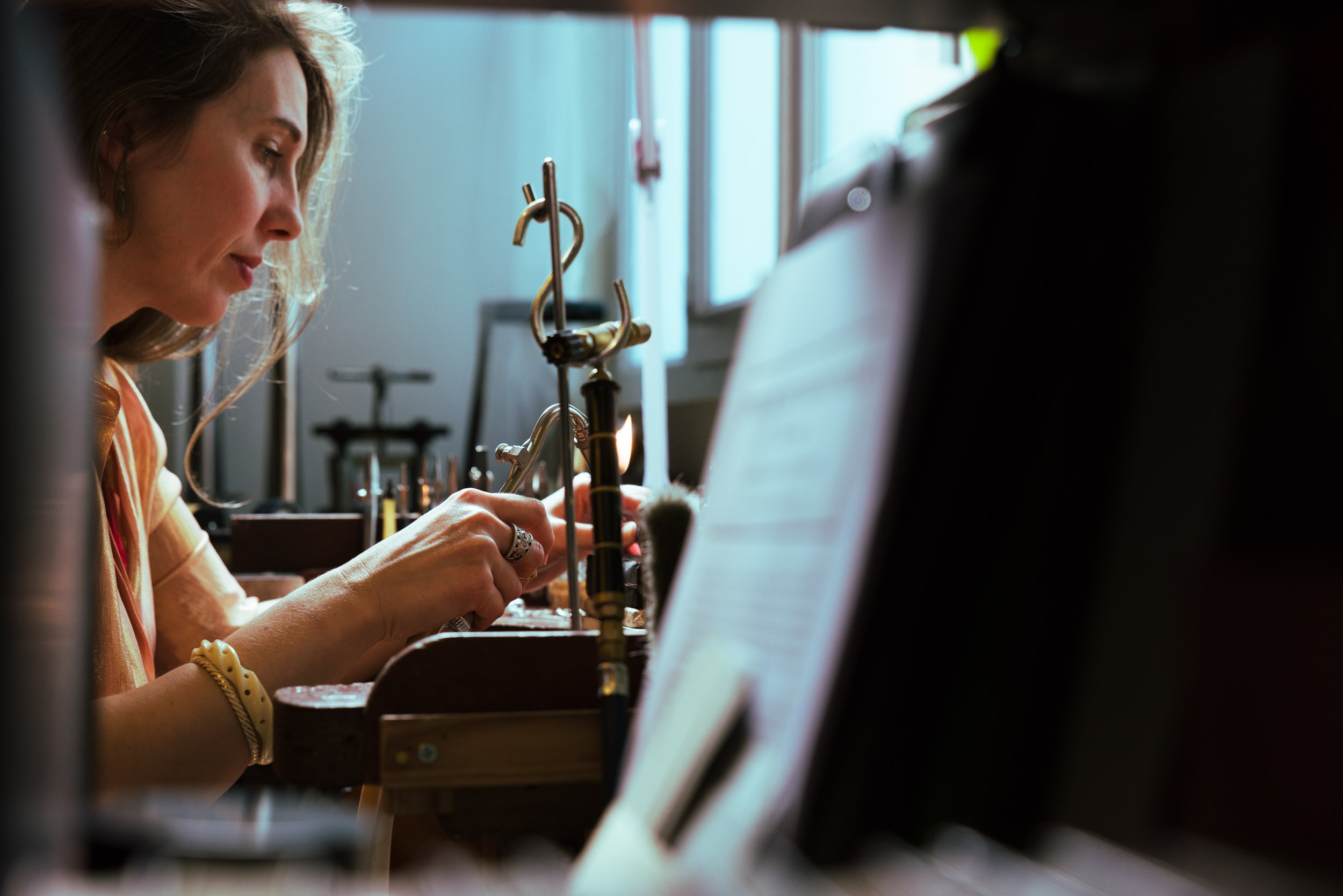 Une femme est assise à un établi, concentrée sur un bijou qu'elle est en train de fabriquer. Entourée de portraits d'artisans de Bordeaux et de Gironde, elle utilise des outils pour travailler délicatement sur le bijou dans son espace de travail bien éclairé avec d'autres équipements visibles en arrière-plan. La scène évoque la concentration et le savoir-faire.