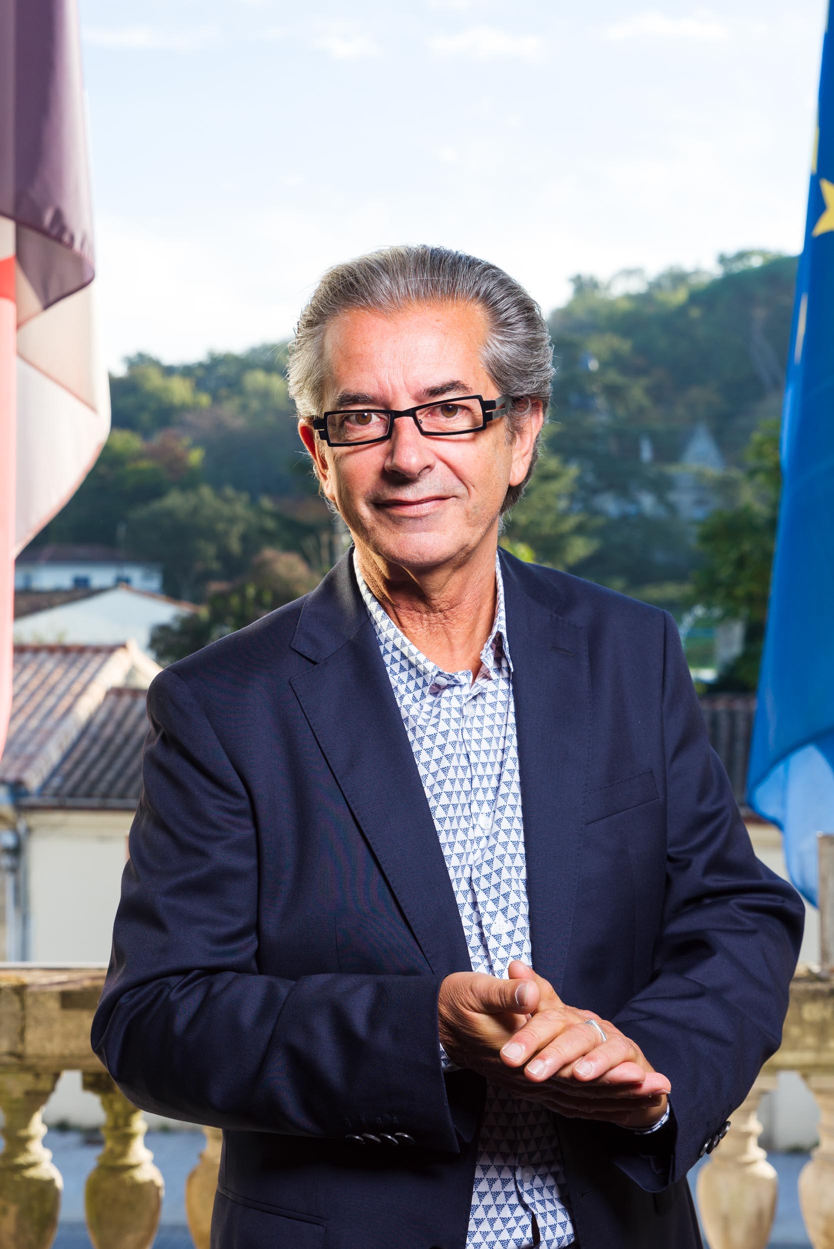 Un homme aux cheveux gris et aux lunettes se tient debout devant une balustrade en pierre. Il porte un blazer bleu foncé sur une chemise à motifs. Derrière lui, on voit des arbres, des toits et des drapeaux flous : l'un rouge et blanc, l'autre bleu avec des étoiles jaunes. Il a un léger sourire et tient ses mains jointes, parfait pour les portraits professionnels à Bordeaux.