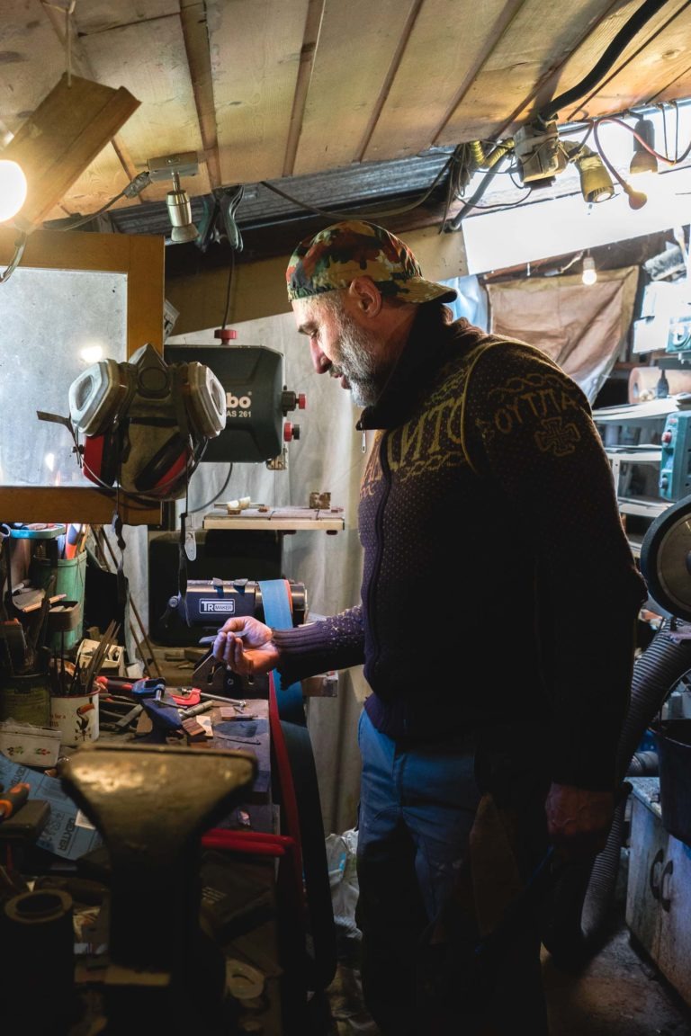 Un homme dans un atelier se tient devant un établi encombré, tenant un objet métallique. Il porte un foulard à motifs, un pull sombre et un jean. La pièce est faiblement éclairée par des poutres en bois apparentes et divers outils éparpillés un peu partout. Prendre des portraits d'artisans comme lui est courant à Bordeaux et en Gironde.