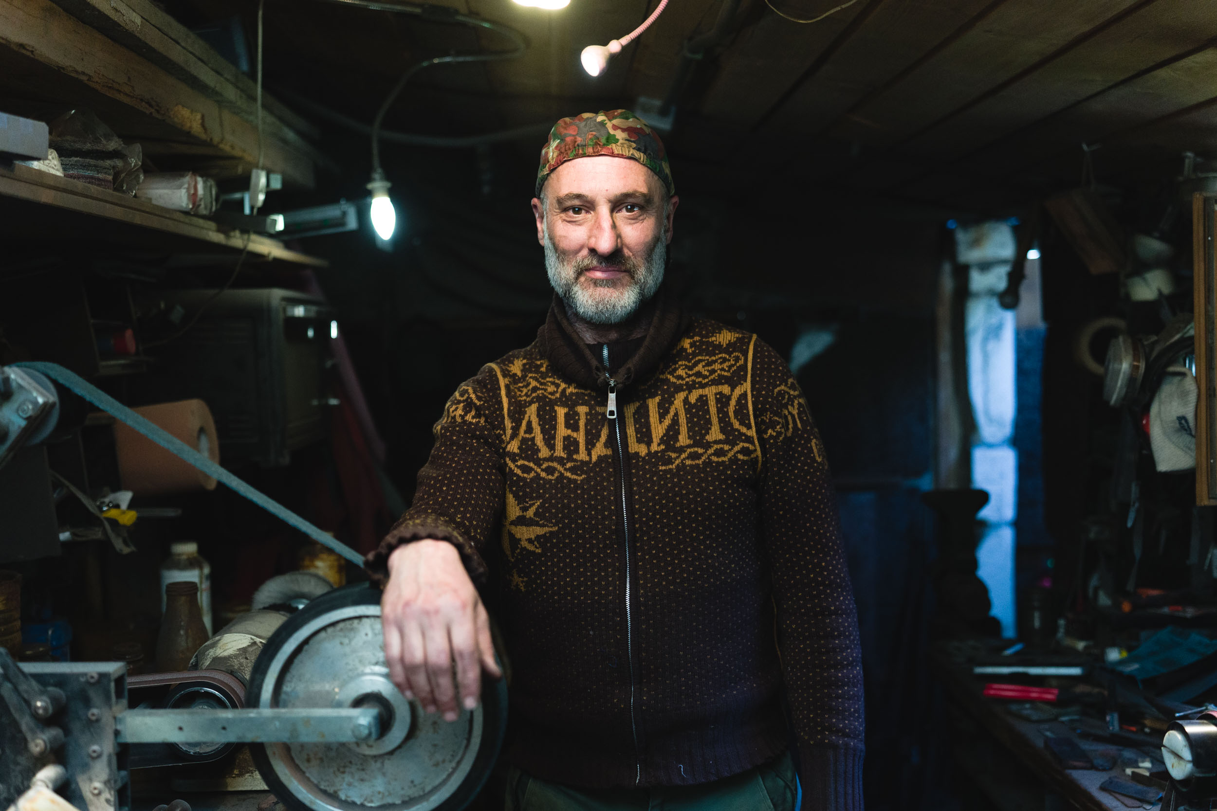 Un homme barbu et moustachu se tient debout dans un atelier faiblement éclairé, son bras droit reposant sur une machine en métal. Vêtu d'un pull marron à motifs et d'une casquette camouflage, il incarne l'essence des artisans que l'on peut rencontrer dans les portraits de Bordeaux et de Gironde. Divers outils et équipements contribuent à l'atmosphère encombrée et industrieuse tandis qu'une ampoule est suspendue au plafond.