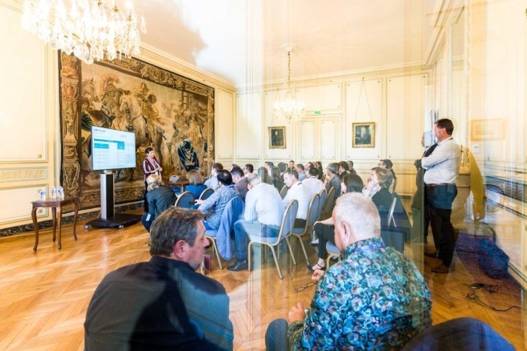 Un groupe de personnes est assis dans une salle spacieuse au décor richement décoré du Palais de la Bourse de Bordeaux, avec notamment des lustres et une grande tapisserie au mur. Ils écoutent attentivement un présentateur près d'un grand écran diffusant une présentation, rappelant une soirée AXA Business. Certains individus se tiennent debout au fond de la salle.