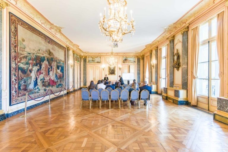 Une salle spacieuse et décorée au Palais de la Bourse de Bordeaux, avec d'élégants lustres et de grandes tapisseries sur les murs. La lumière du soleil entre par de hautes fenêtres avec des rideaux. Un petit groupe de personnes est assis sur des chaises bleues et dorées disposées en rangées, face à un écran blanc à l'avant, probablement pour une soirée AXA Business. Le sol est doté d'un parquet en bois complexe.