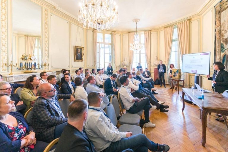 Un groupe de personnes en tenue professionnelle sont assises dans une salle élégante et décorée du Palais de la Bourse de Bordeaux, avec des lustres et de grandes fenêtres. Ils font face à un conférencier qui présente des diapositives sur un grand écran, faisant de cette soirée un événement AXA Business sophistiqué, avec des murs couleur crème, des tableaux encadrés et des rideaux allant du sol au plafond.