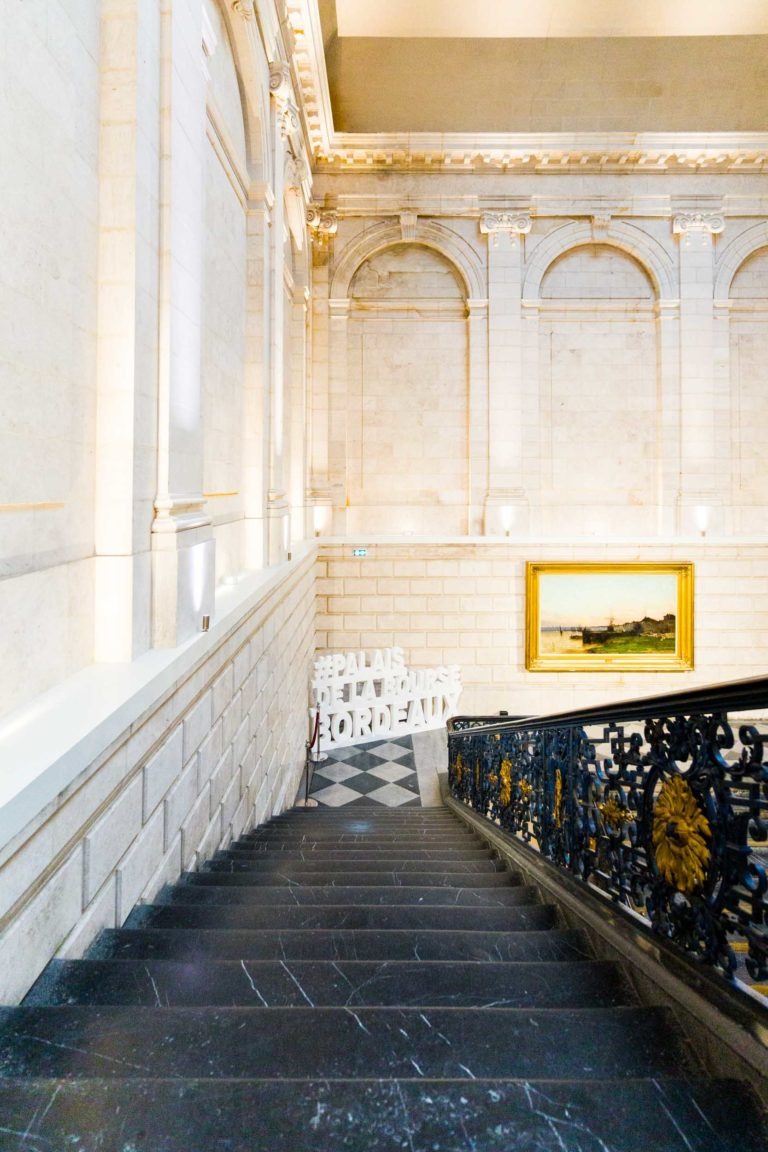 Un grand escalier aux marches en marbre noir et aux rampes noires et dorées ouvragées mène à un grand mur de pierre décoré d'un seul tableau de paysage encadré. À gauche, une installation lumineuse énonce "#PALAS #EXPLAMES #JORDENAV", ajoutant une atmosphère de soirée rappelant l'élégance du Palais de la Bourse à Bordeaux.